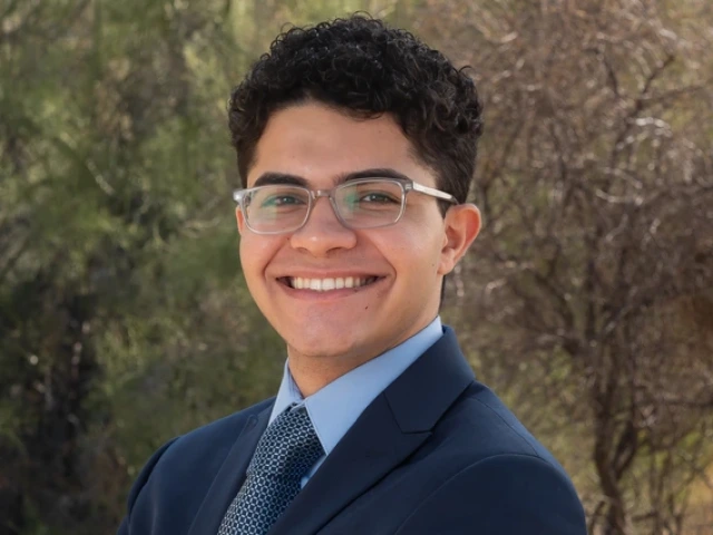 A young man with short curly hair and glasses smiles confidently in a professional outdoor portrait. He wears a navy blue suit, a light blue dress shirt, and a patterned tie, standing against a natural desert background with green and brown foliage.
